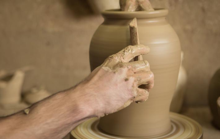 Imagen de una pieza de arcilla que va tomando forma, junto a las manos expertas de un alfarero, que la modela al torno con la ayuda de una caña para alisar la superficie.